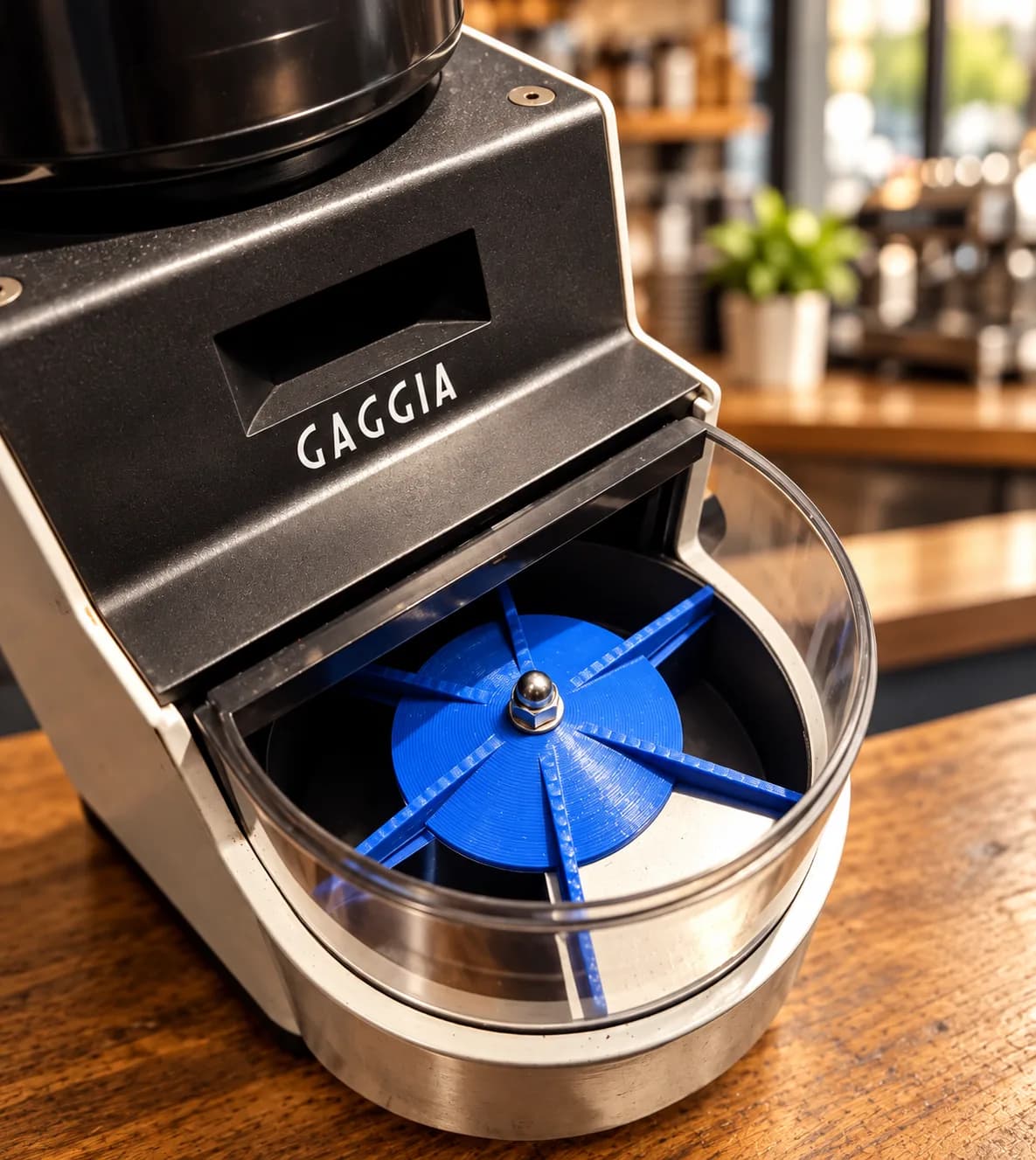 A blue 3D printed doser impeller fitted inside a Gaggia MDF grinder's doser, lid open, on a wooden café counter