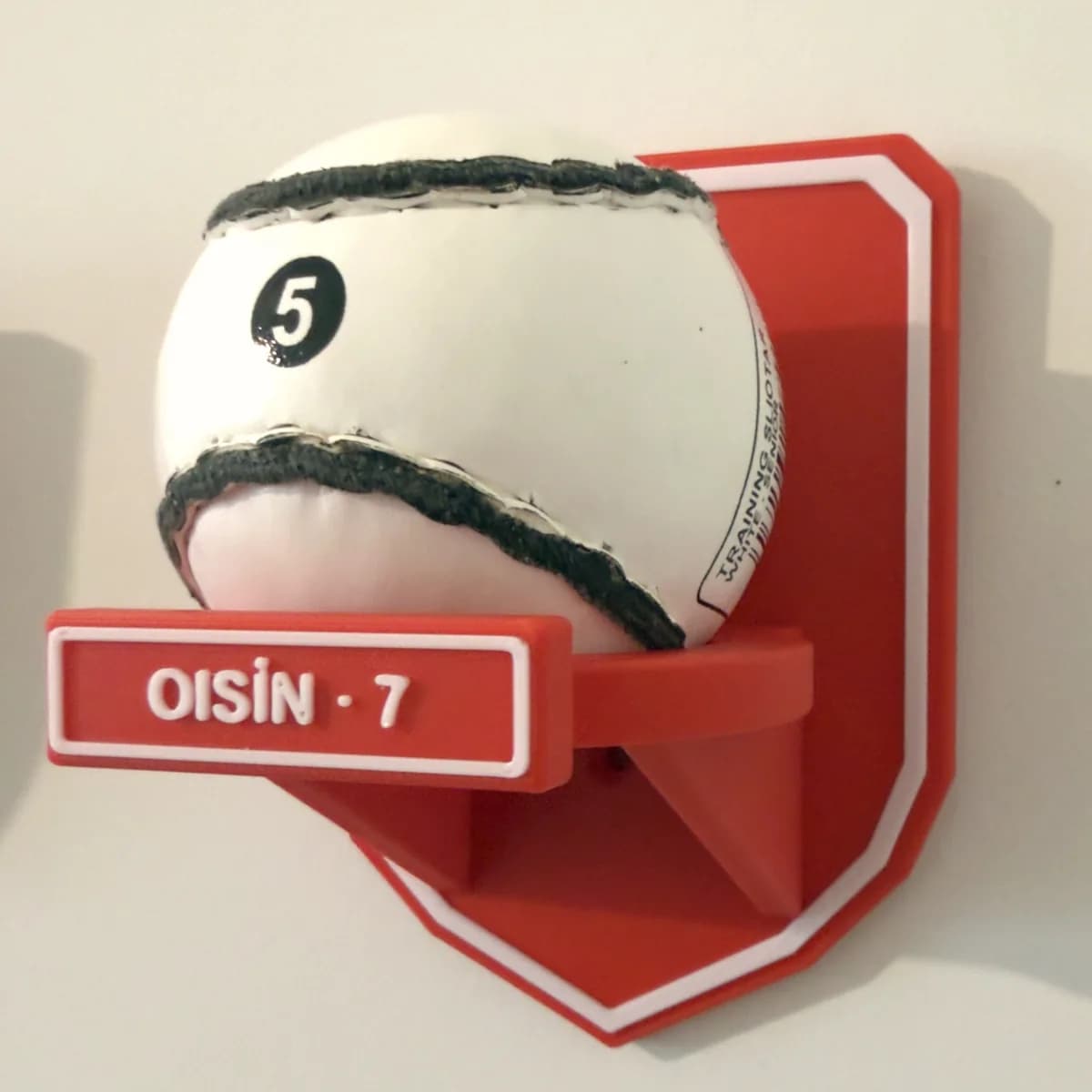 The printed red and white sliotar wall mount holding a sliotar on its open ring, with OISÍN · 7 raised on the name plate